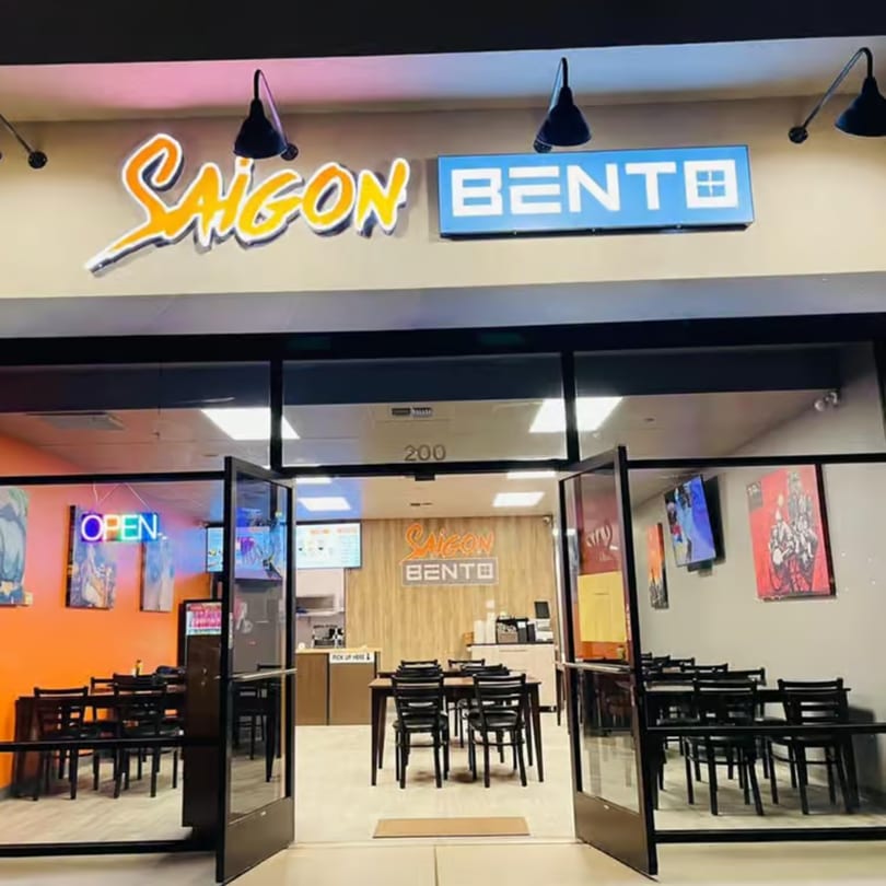 Exterior view of Saigon Bento restaurant at night, with glowing signs and visible interior seating.