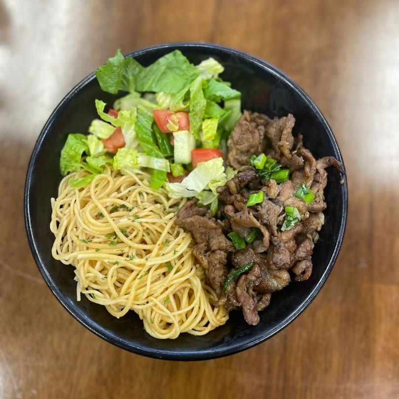 A black bowl of Saigon Bento's Vietnamese noodles, beef, and fresh salad on a wooden table.