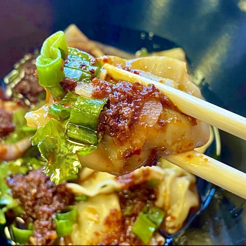 Chopsticks holding a Vietnamese dumpling topped with chili oil and green onions.