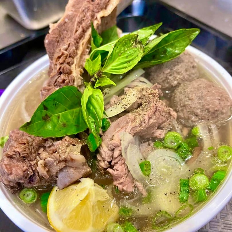 Vietnamese pho with beef, meatballs, basil, onions, and a lemon wedge in a white bowl.
